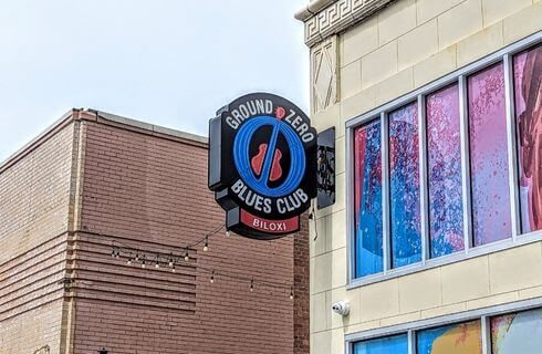 A portion of a brick building with blue, pink and purple colorful windows, with a round sign that says Ground Zero Blues Club