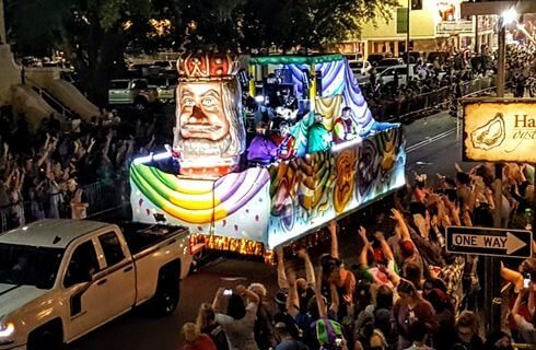 A street with a colorful mardi gras float and hundreds of people lining the street waving at the float and enjoying themselves