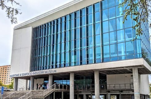 A multi story building with full windows reflecting the blue sky with the words Maritime & Seafood Industry Museum on the front of the building above the entrance