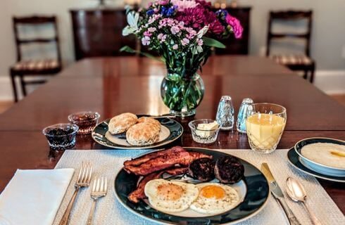 A table set for breakfast with a plate of sunny side up eggs, sausage, jam, bacon, biscuits, a glass of orange juice, and a vase of colorful purple, pink and whie flowers.