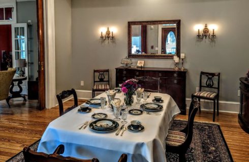 A dining room with wood floors, a tapestry carpet, a table covered with a white table cloth, set with black and white china and silverware, antique furniture along the walls, and an opening to the living room adjoining the dining room.