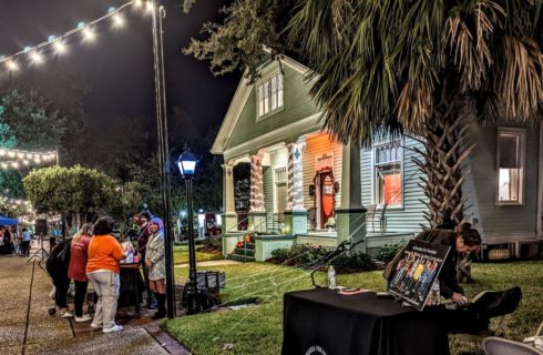 people gathering around a table of food at night in front of a green house light up with lights, palm trees, and hanging lights criss crossed over the street.