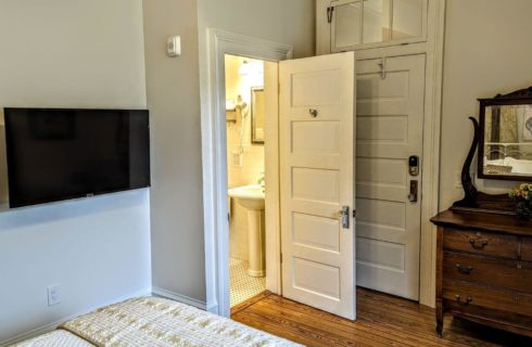 A bedroom with wood floors, the corner of a bed, a flat screen TV on the wall, an antique dresser with mirror against one of the walls, and a door leading into a bathroom.