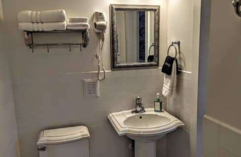 A bathroom with white walls, a pedestal sink, chrome fixtures, a pewter mirror above the sink, a chrome towel rack with white towels, and a hair dyer.