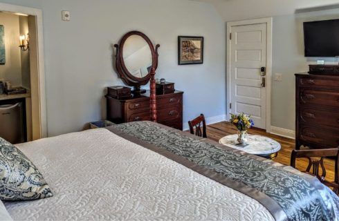 A bedroom with wood floors, a bed with white linens a grey satin and tapestry throw at the foot of the bed, and black and cream colored throw pillow, a small marble table and 2 chairs at the foot of the bed, dark antique dressers against the walls, a flat screen TV on the wall, and a doorway to a small closet with a small refrigerator and coffeemaker.