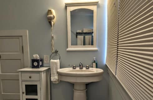 A bathroom with grey walls, white trim, a pedestal sink with chrome fixtures, a mirror above the sink, a white cupboard to the side of the sink.