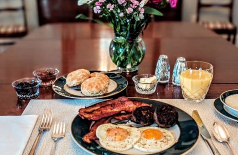A wood table set for breakfast with a black and white plate of sunny side up eggs, sausages, bacon, and side bowl of grits, a side plate of biscuits with bowls of jam and butter, a glass of orange juice, and a vase of pink flowers.