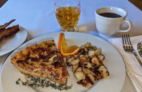 A white plate with a piece of quiche, roasted potatoes, garnished with a slice of orange and sprig of thyme, with silverware on a cloth napkin next to it, another plate with crisp slices of bacon, a crystal glass with juice, and a white cup with black coffee.