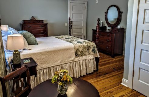A bedroom with grey walls, white trim, wood floors, a bed with a white bedspread and green and cream colored throw a the foot of the bed, green accent pillow, a wood nightstand with lamp next to the bed, a wooden dresser with mirror and chest of drawers against the walls, and a small wood table and chair .