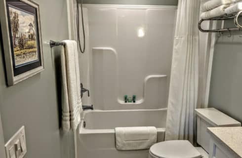 A bathroom with grey walls, white trim, a tub and shower combination, toiletries on a shelf above the tub, a toilet, sink, a bath mat on the tub, and white towels hanging on a rack next to the tub.