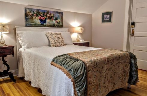 A bedroom with wood floors, a bed with white bedspread and pillowcases, a tapestry throw at the foot of the bed, a wood nightstand with a lamp next to either side of the bed, and a picture of potted flowers above the bed.