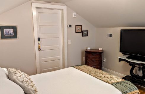 A bedroom with a bed with white bedspread and pillowcases, a tapestry throw at the foot of the bed, a flat screen TV on a wood and marble stand, a small dresser in the corner, and a lamp next to the bed.