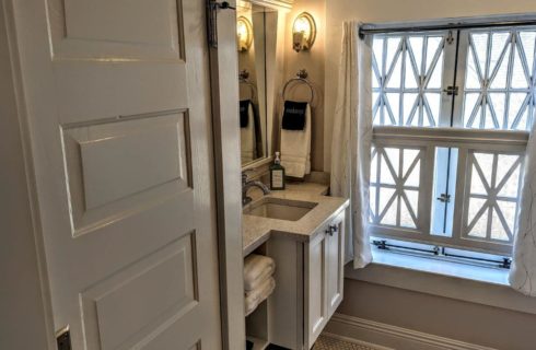 A bathroom with grey walls, white trim, a white door, a single sink with chrome fixtures, white towels, and a window letting in plenty of light.