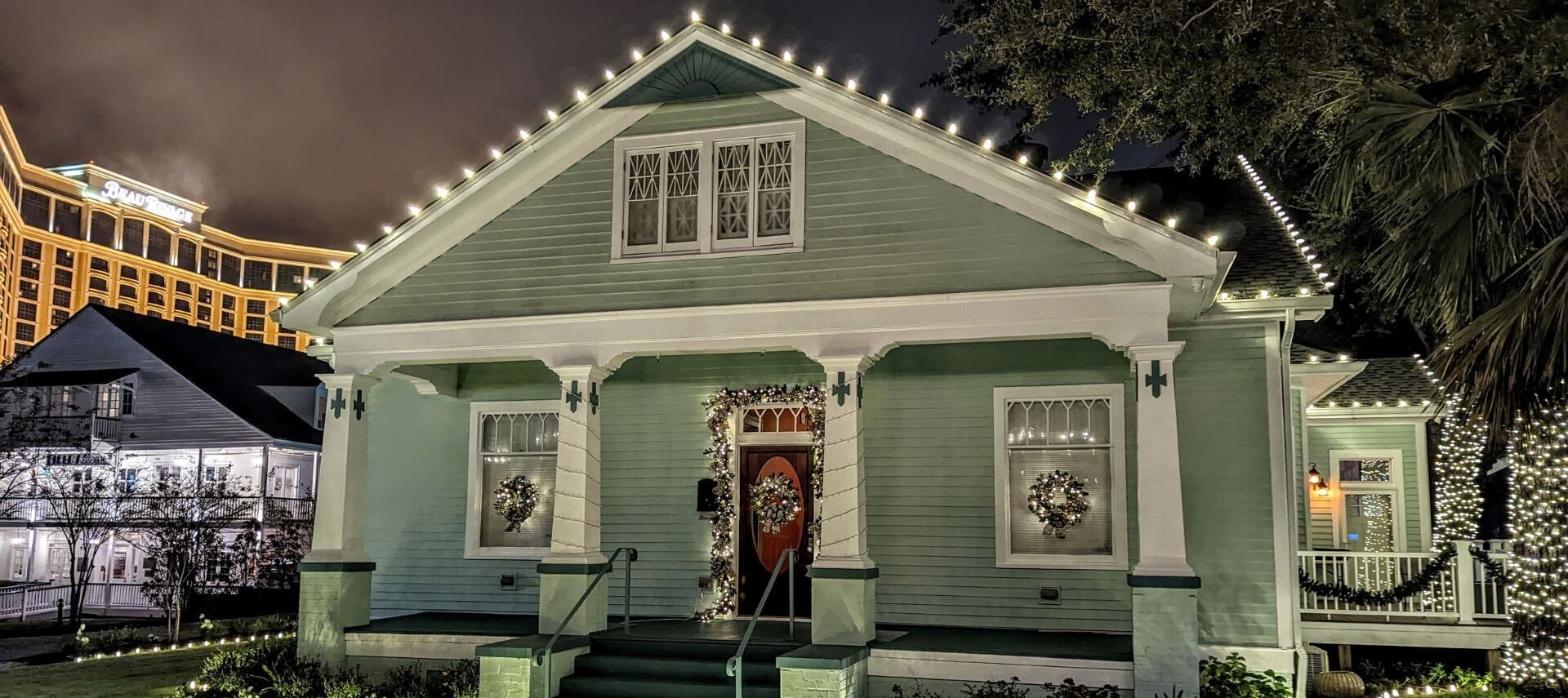 A green 2-story Victorian house with white trim and pillars, light up with Christmas lights.