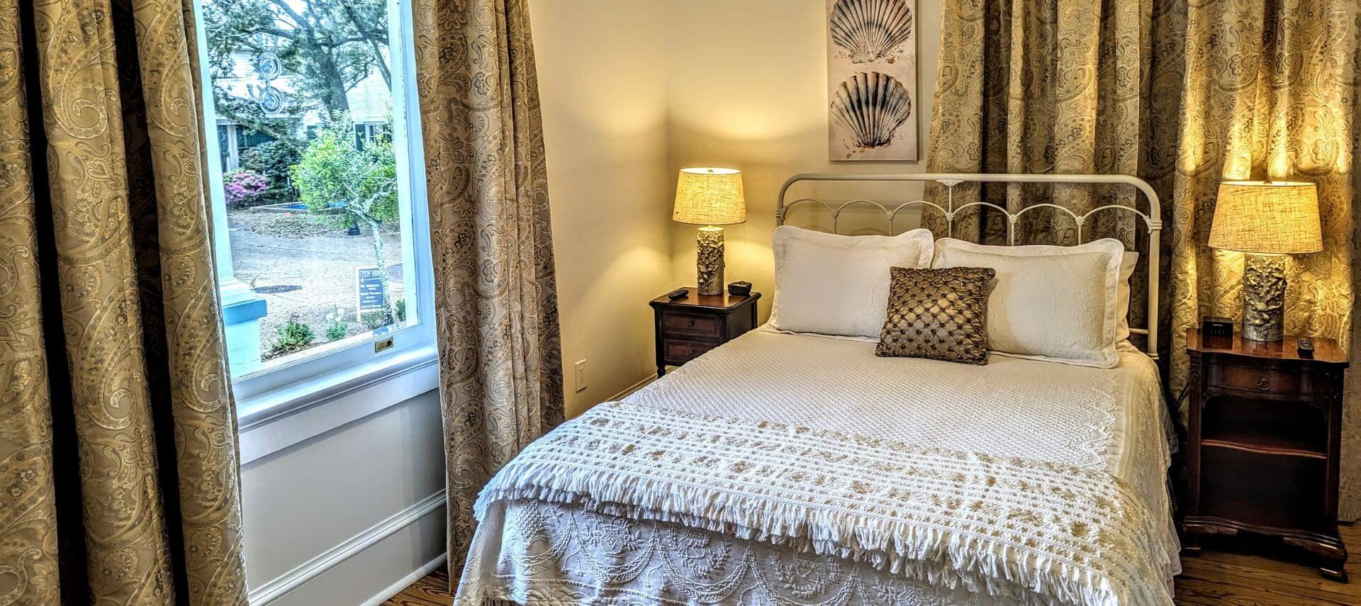 A bedroom with a bed with wrought iron headboard, white bedding, seashell photos above the the bed, a nightstand with lamp next to the bed, and a window letting in light with tapestry curtains.