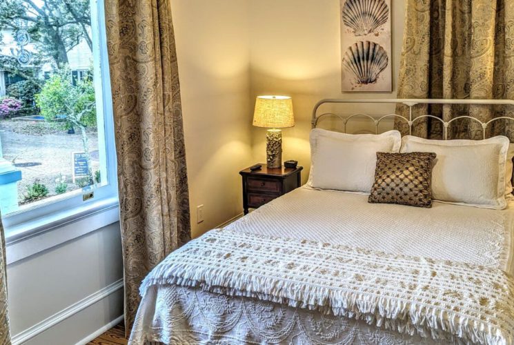 A bedroom with a bed with wrought iron headboard, white bedding, seashell photos above the the bed, a nightstand with lamp next to the bed, and a window letting in light with tapestry curtains.