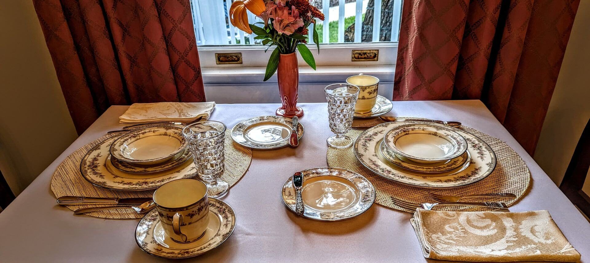 An elegantly set table with china, crystal glassware, silver ware, a vase of fresh flowers, and a window letting in sunlight with a view of the outside greenery