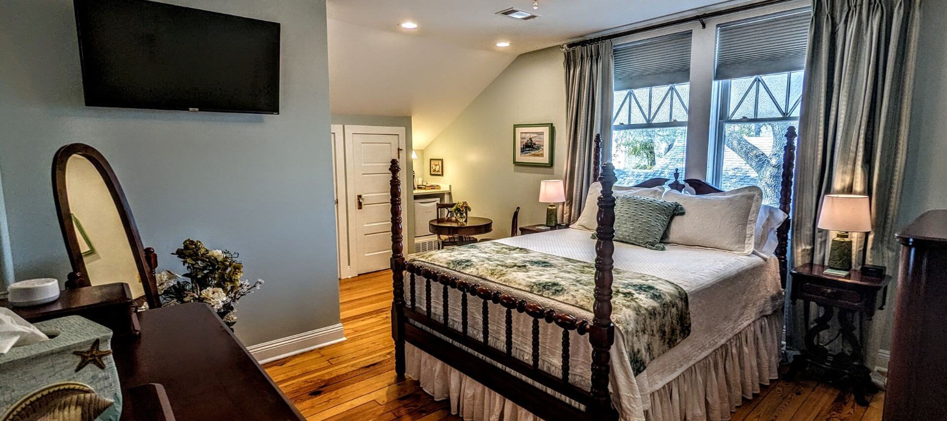 A bedroom with cream and sage colored walls, wood floors, a 4 poster bed with white bedding and a colorful quilt, with windows and green curtains, along with a full length oval mirror and flat screen TV
