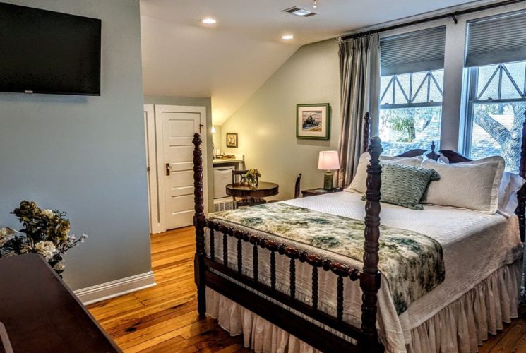 A bedroom with cream and sage colored walls, wood floors, a 4 poster bed with white bedding and a colorful quilt, with windows and green curtains, along with a full length oval mirror and flat screen TV