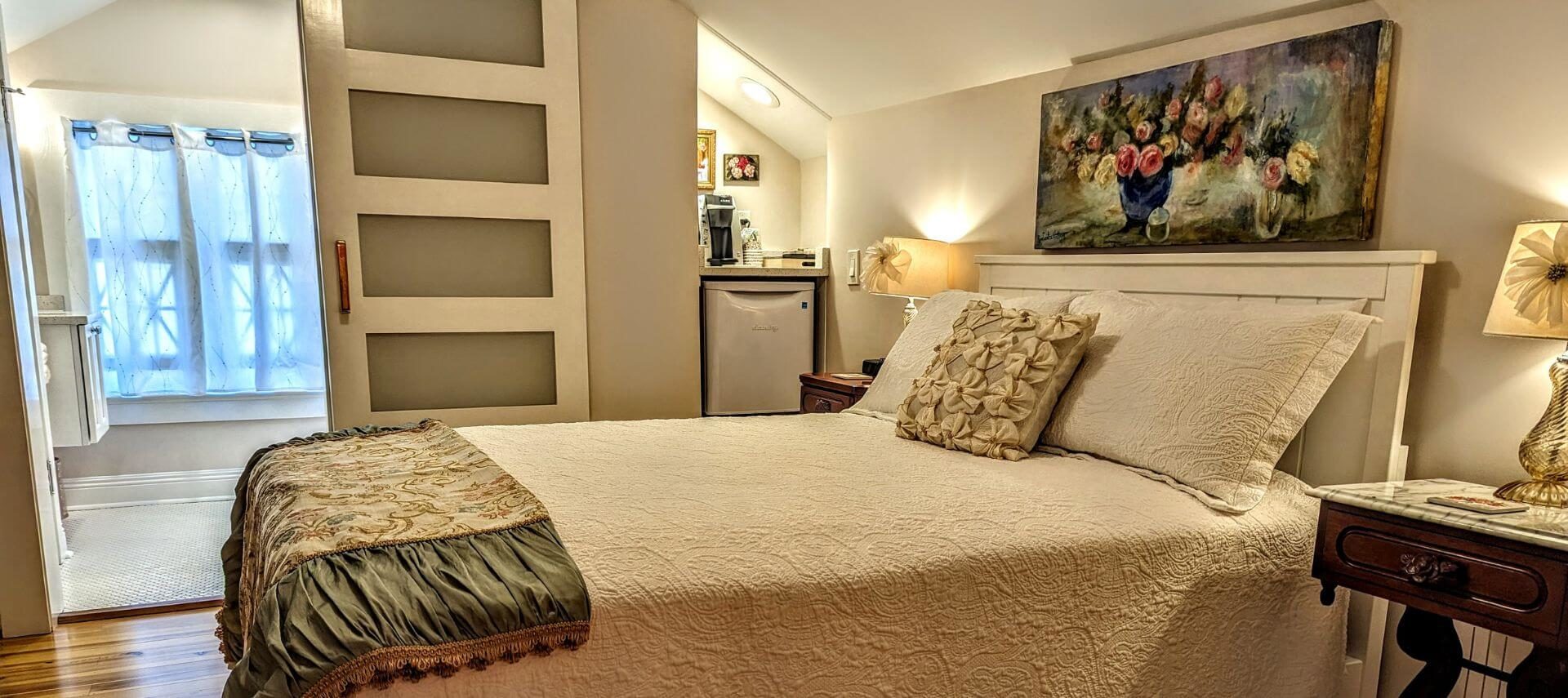 A bedroom with wood floors, cream and green walls, a bed with white bedding and tapestry throw, a framed photo above the bed, a mini fridge and coffee center, and a window letting in sunlight