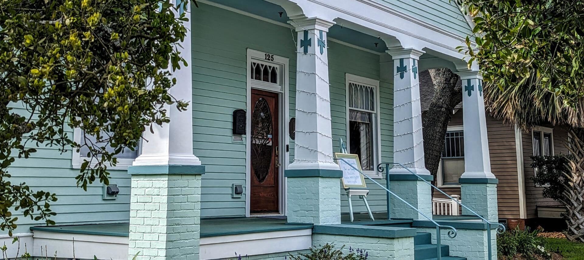 A light green Victorian front porch with white pillars and green bushes