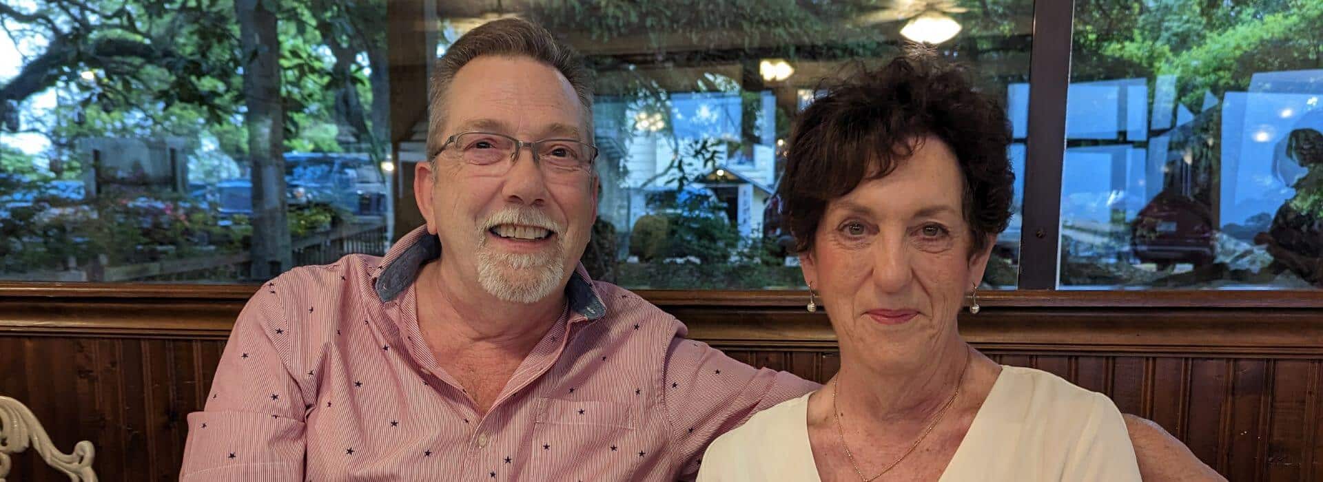 a smiling man with a salmon colored shirt, a beard and glasses, with his arm around a smiling woman with dark hair, wearing a yellow shirt and earrings.