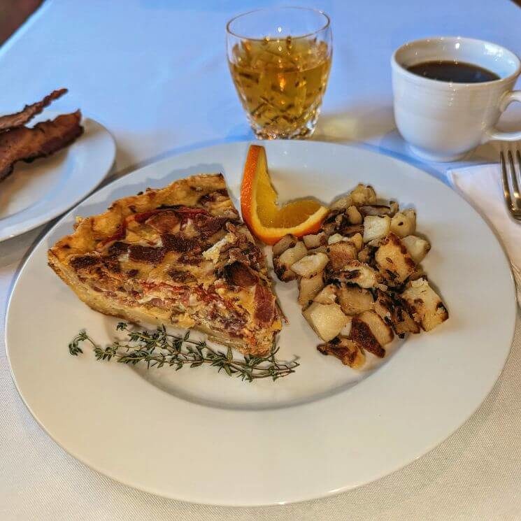 A table set for beakfast with a quiche, potatoes and garnish on a white plate, along with a side of bacon, a glass of juice, and a cup of coffee.