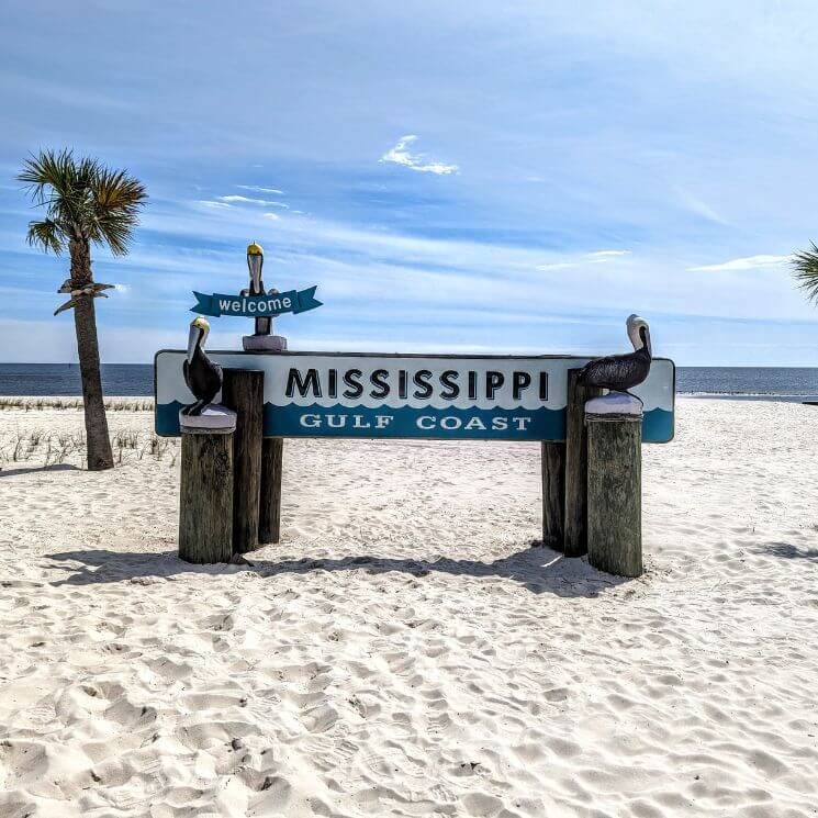 A blue and white sign that says MISSISSIPPI Gulf Coast on wood pillars on a sandy beach with palm trees on either side