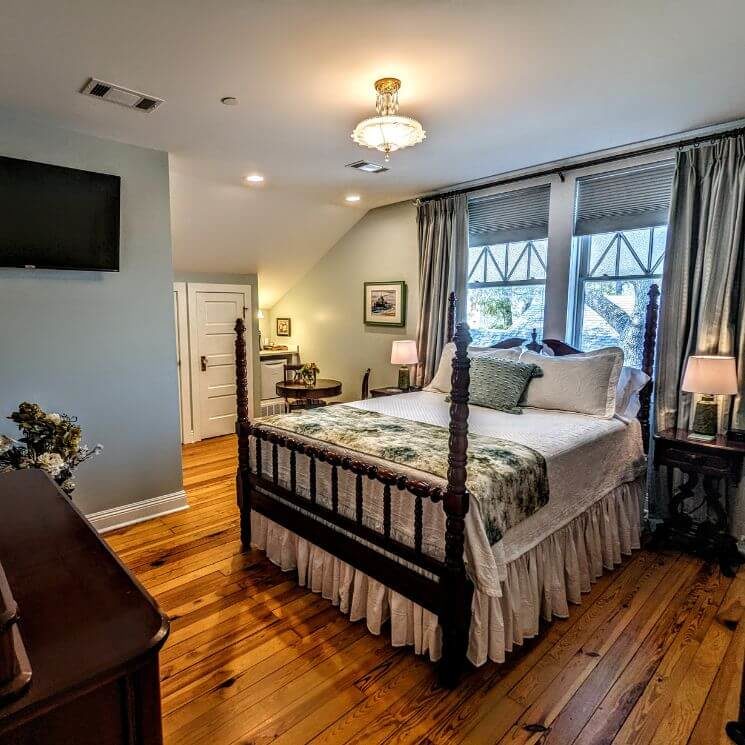 Picture of a bedroom with a fourposter bed, white bedding, tapestry throw and pillow, wood floors, a flat screen TV on the wall, and windows with green curtains