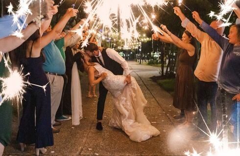 A bride and groom kissing on a street with well wishers on either side of them holding up sparklers.